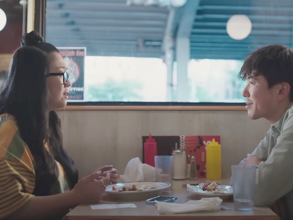 Two people sit across from each other at a table in a diner. On the left, an Asian woman with long dark hair and glasses sits with her hands held up over her plate, mid-speech. On the right, an Asian man with short dark hair and a grey button-up shirt leans forward with his elbows on the table, smiling at her.