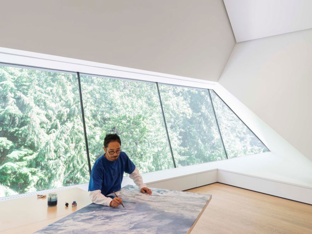 A man wearing a blue T-shirt over a white longsleeve is painting on a large canvas on a table. The room is uniquely angled, with large panelled windows showing lush forest.