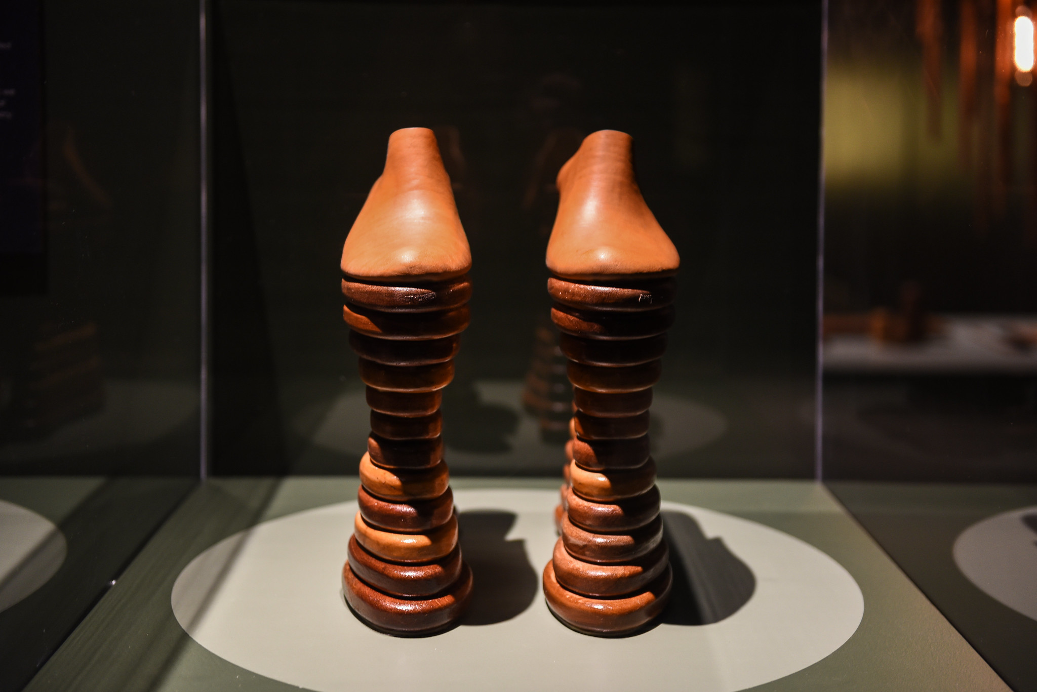 A pair of mahogany shoes stands in a glass gallery box atop a smooth stack of round pieces of glossy milled wood.