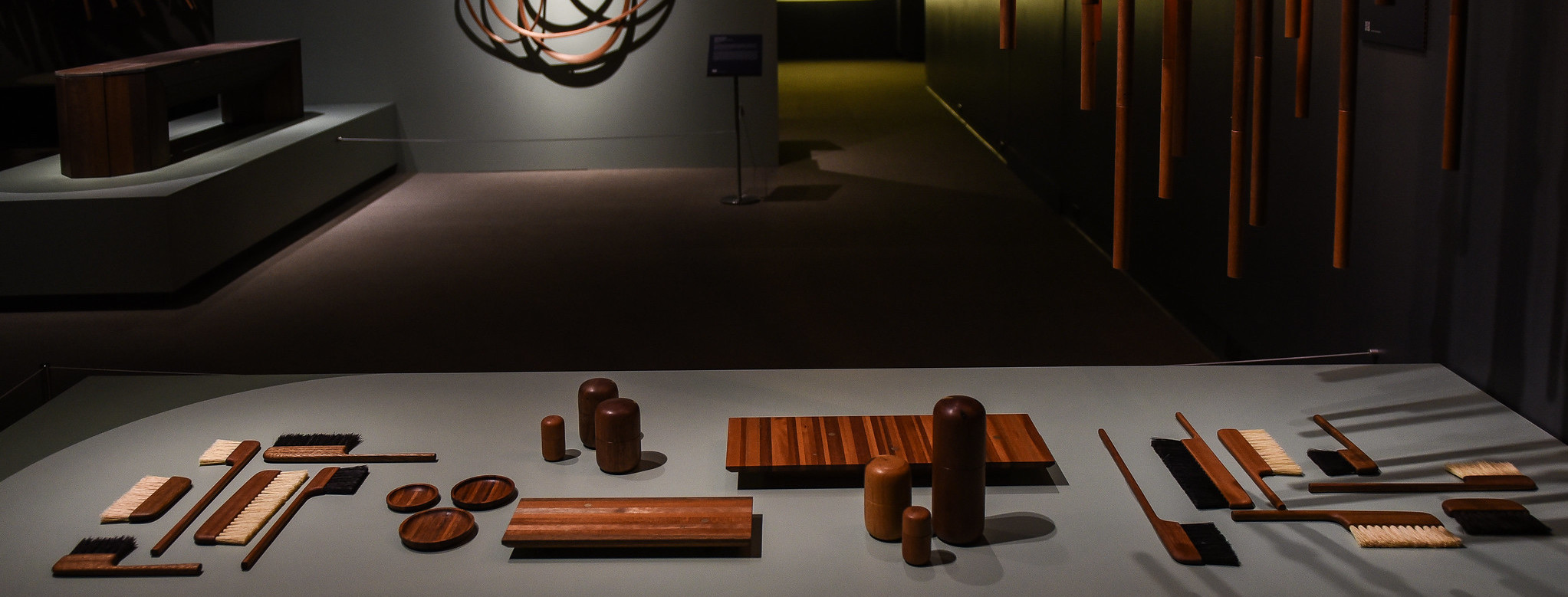 A range of mahogany objects sits across a low grey table in a gallery space with grey walls and soft lighting. The objects include brushes, coasters and cylindrical objects. In the background are abstract mahogany wall sculptures and, to the left in the background, a bench.