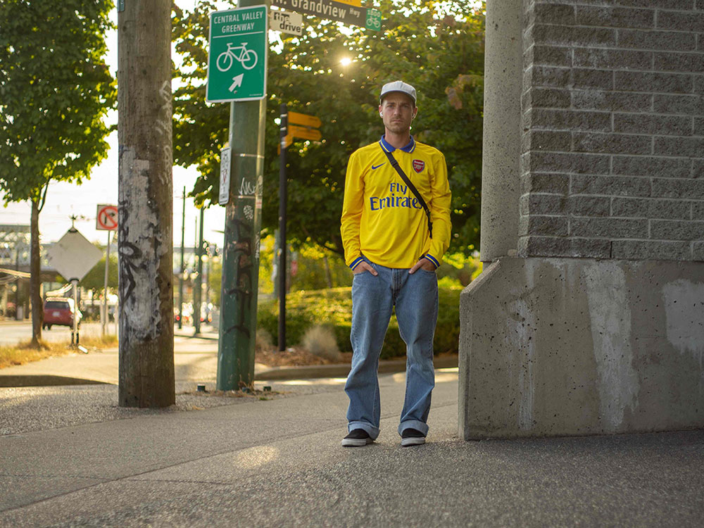 Mike DiPietro is wearing a white ball cap and yellow long-sleeved soccer jersey, light wash jeans and black skateboarding shoes. He is standing on a sidewalk near Grandview Highway in East Vancouver. Behind him is a stand of sun-dappled deciduous trees.