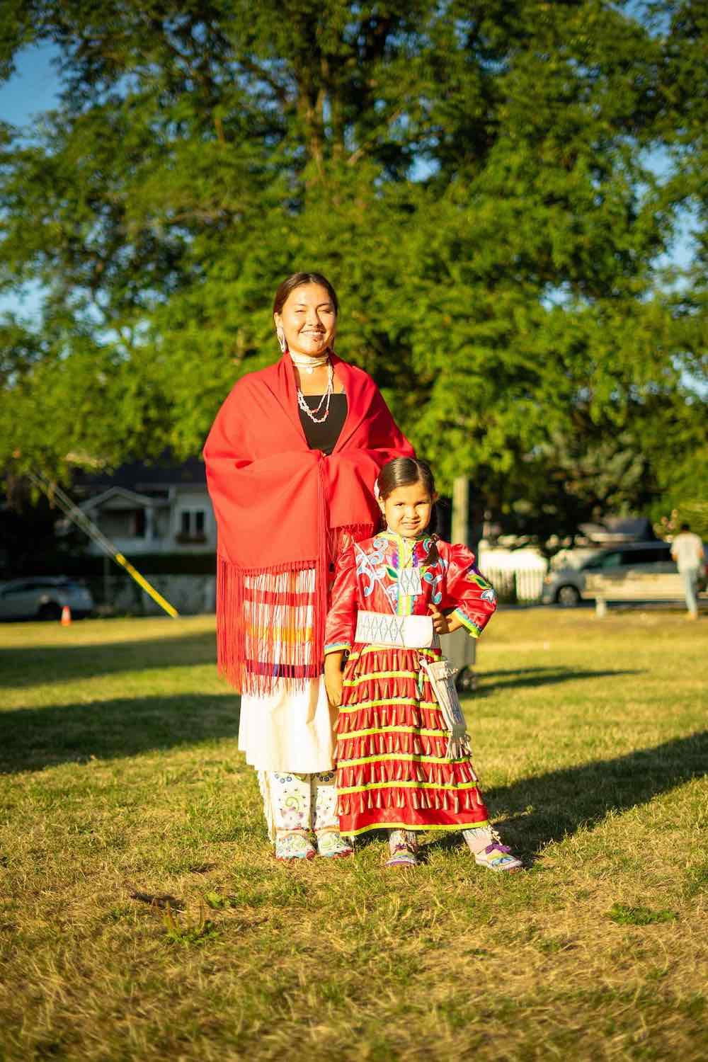 A woman and child stand together in the grass in a sunny neighbourhood park. They are wearing festive red Indigenous regalia in bright red. They are smiling at the camera. 