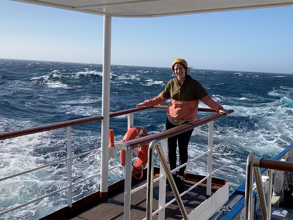 Laura Trethwey stands near the railing of an upper deck of Nautilus. The sea is rough behind her.