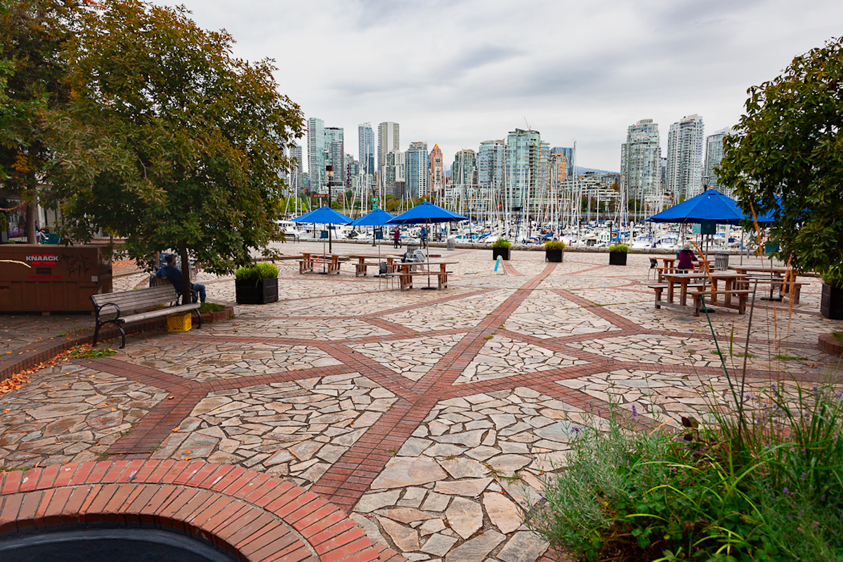 A paving stone courtyard with straight red brick lines embedded in the stones and pointing at different angles towards the waterfront. There are sailboats and yachts in the water and condo towers on the horizon.