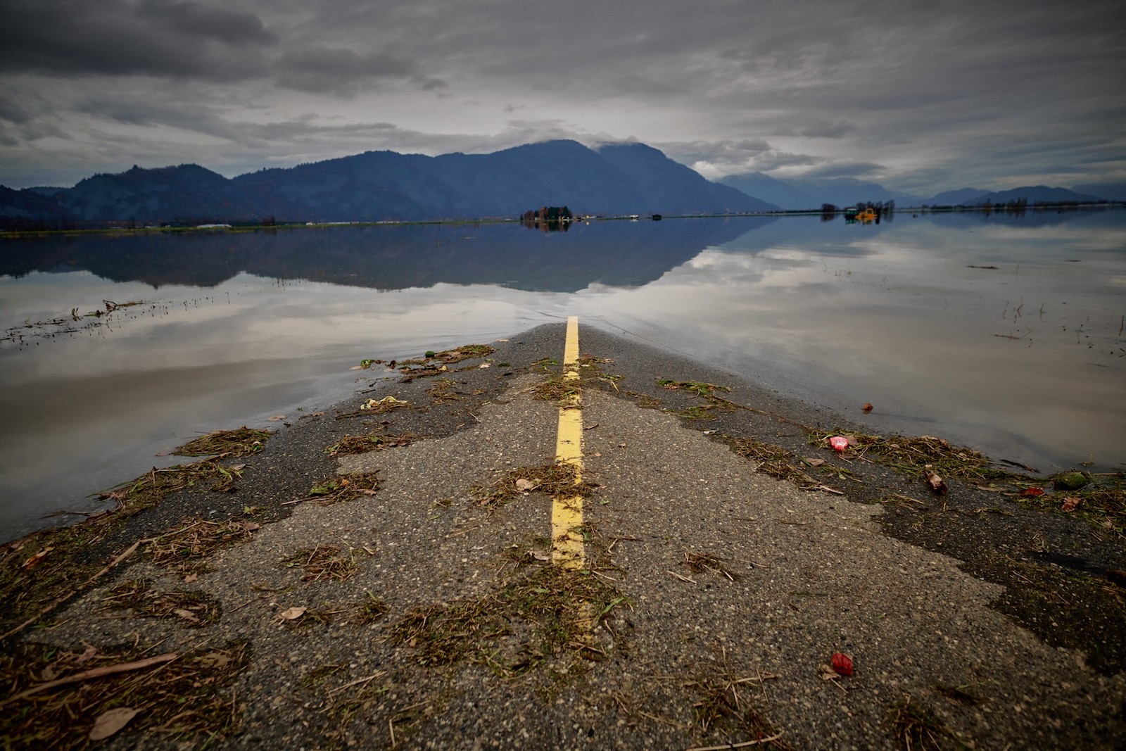 A single yellow line highway with debris in the foreground is covered by floodwaters in the mid-ground, with mountains and cloudy skies in the background.