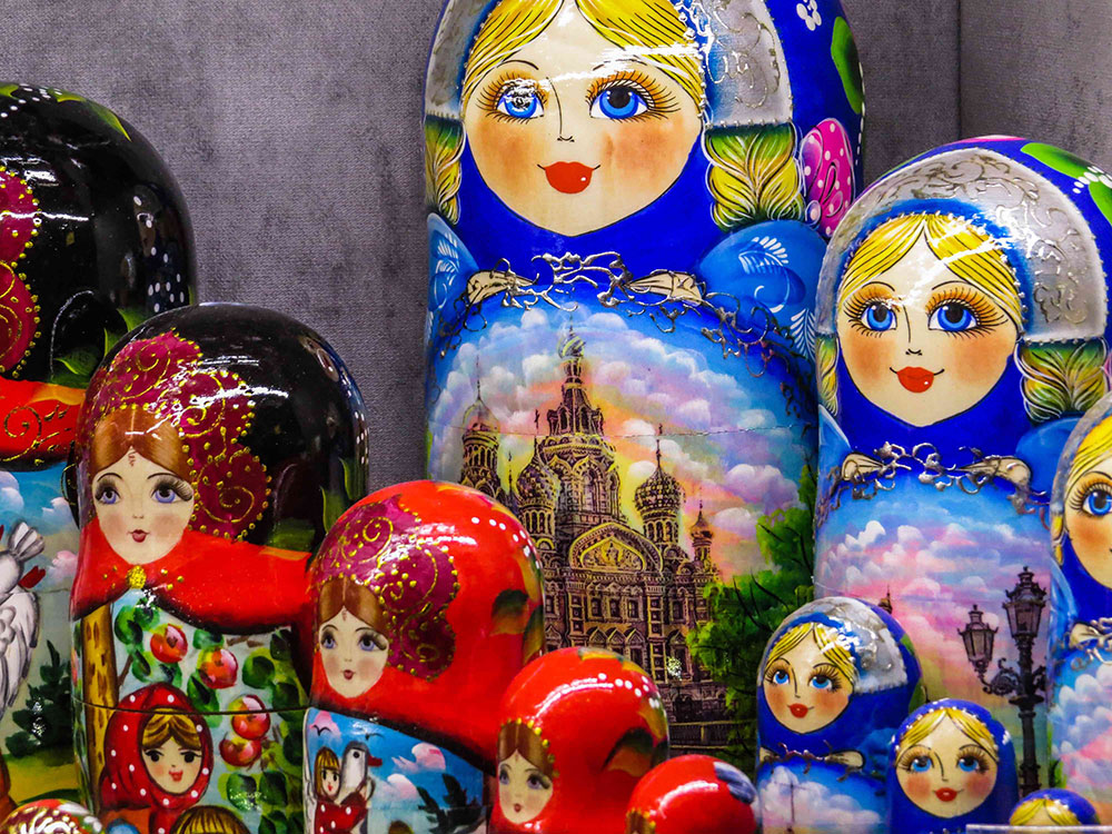 An assortment of traditional Russian matryoshkas (nesting dolls) stand next to each other in a close-up photograph. Blue dolls with blond hair form the focal point of the shot; in the foreground stands a collection of red dolls with brown hair.
