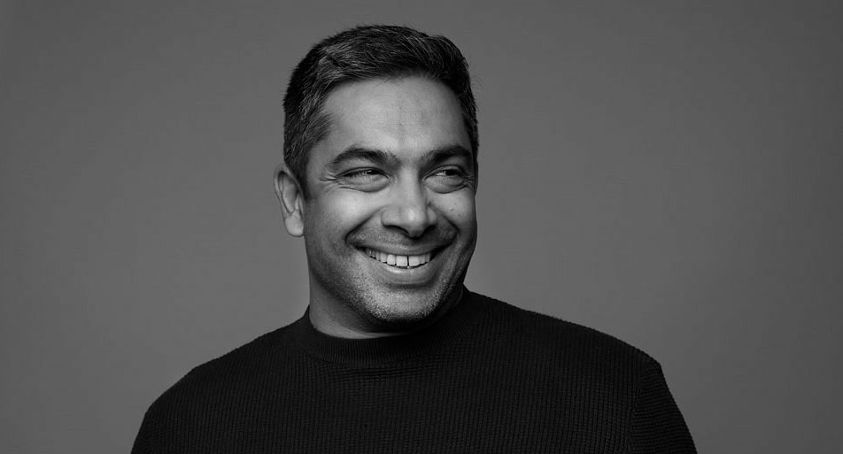 A black and white headshot of Sirish Rao features Rao smiling and looking towards the right of the frame. He is wearing a black crewneck shirt.
