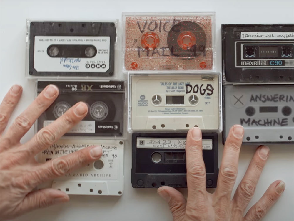 Two hands arrange a collection of black, white and grey cassette tapes on a white surface. Each tape is labelled in handwriting using a black permanent marker.