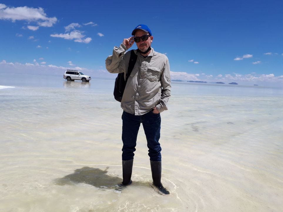 A man stands ankle deep in water over a salt flat, with a blue sky above, low hills in the distance and a white SUV visible in the background. He is wearing a blue cap, sunglasses, a black medical mask under his chin, a tan long-sleeved collared shirt, jeans and rubber boots.
