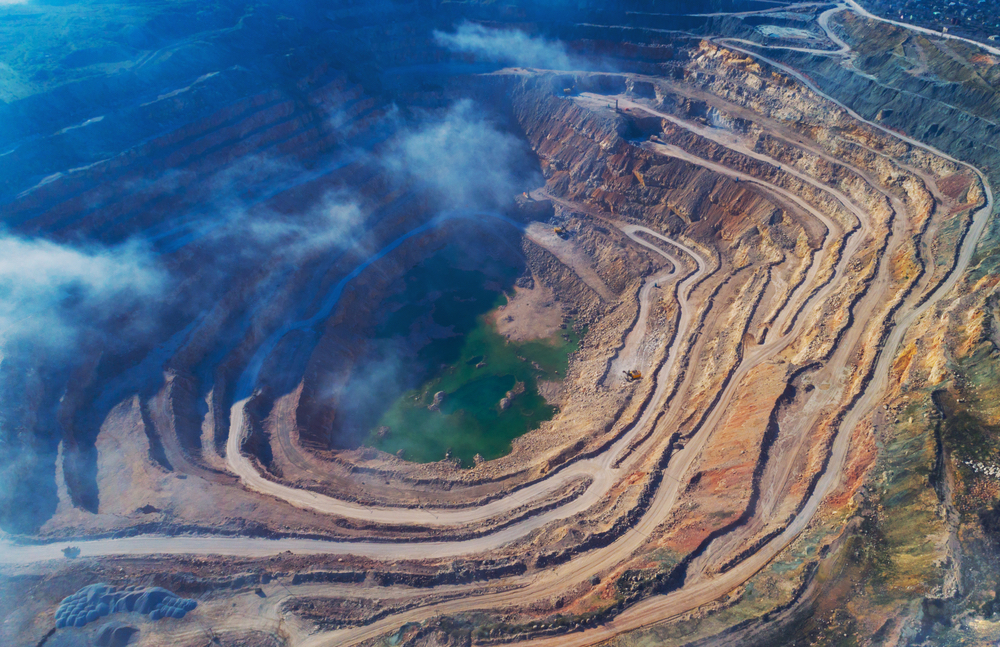 An aerial view of an open pit mine shows a green lagoon at the bottom surrounded by concentric rings of bare dirt ledges/roads.