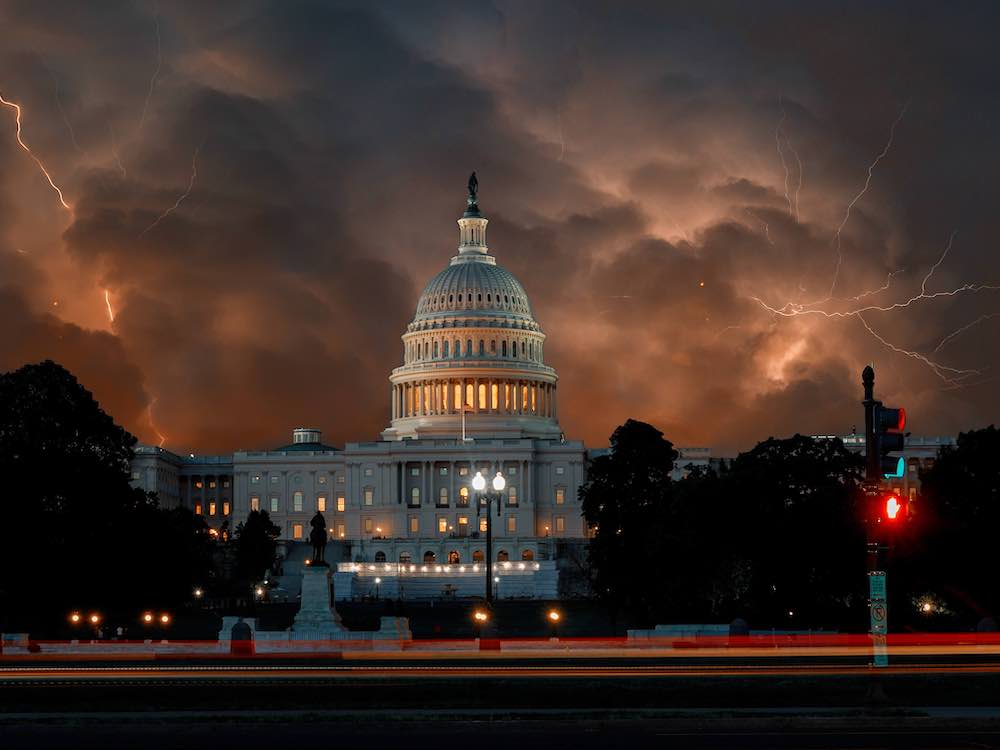 Indoor lights are visible in the United States Capitol at night. The tall central cupola is flanked by dark crops of trees and shrubbery. A stormy, cloudy sky looms above. Lightning bolts are visible amidst the orange and grey.