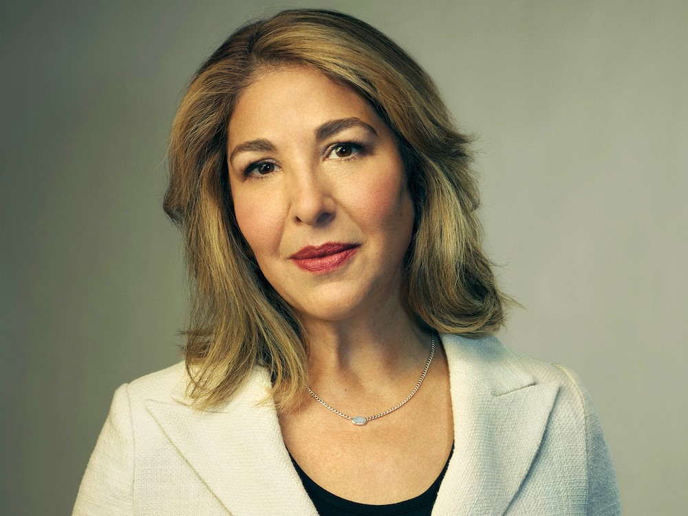 Naomi Klein looks directly at the camera against a grey studio backdrop. She has shoulder-length brown hair with blond highlights. She’s wearing a white linen blazer, a silver necklace and a black top.