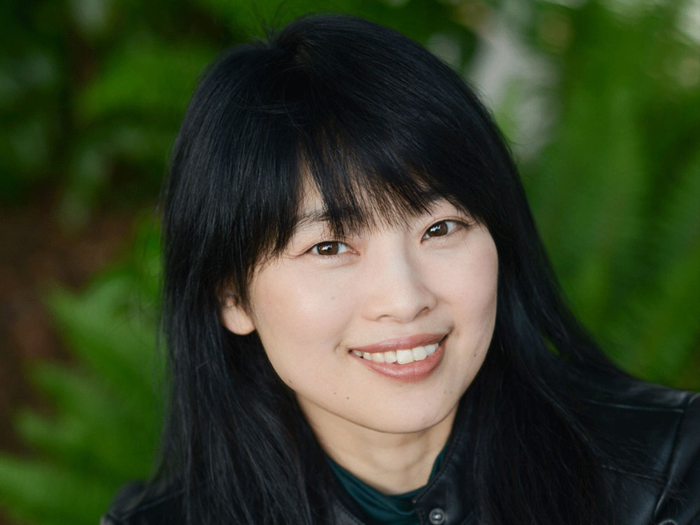 Close-up of Keiko Honda against a soft-focus background of lush green. She has long black hair with bangs. She is looking directly at the camera, smiling.