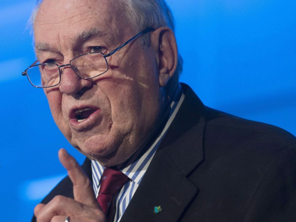 Close-up of Ed Broadbent shows a man with grey hair and glasses speaking while holding up his index finger. He is wearing a black jacket, a blue and white striped shirt and a burgundy-coloured tie.