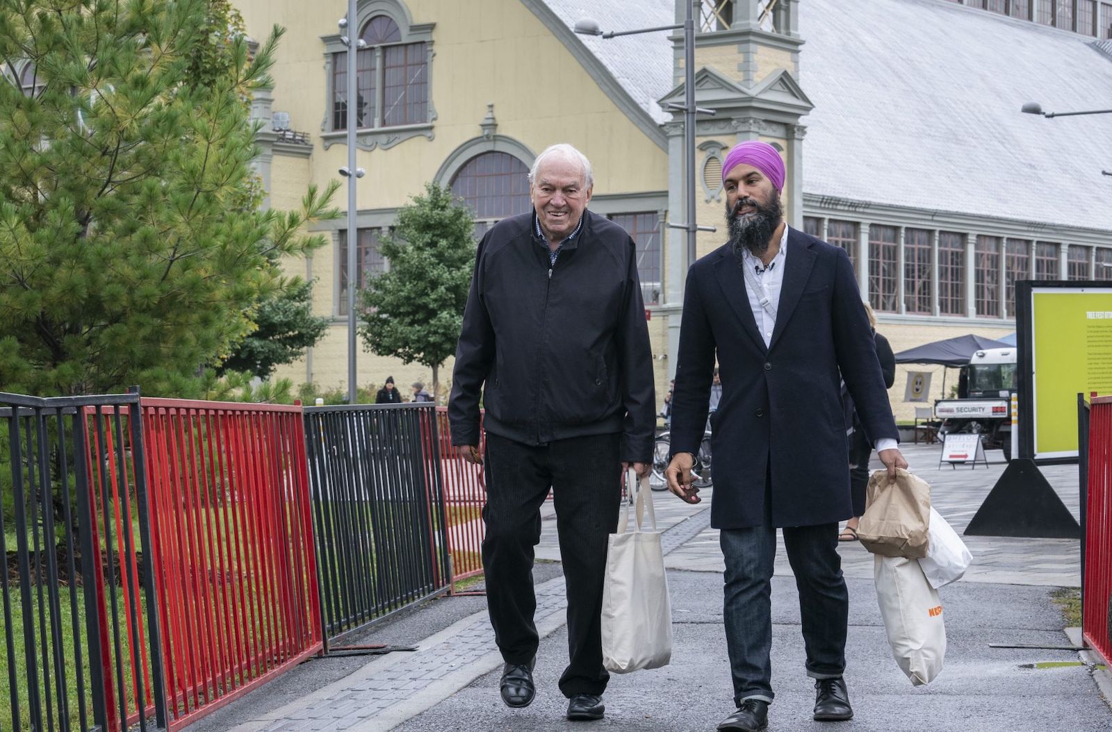 Ed Broadbent, on the left, with thinning white hair, dressed all in black and carrying a canvas shopping bag, and Jagmeet Singh, bearded, wearing a purple turban, a long black coat and jeans and carrying three shopping bags in one hand, walk side by side in front of a large beige building with multiple paned windows.