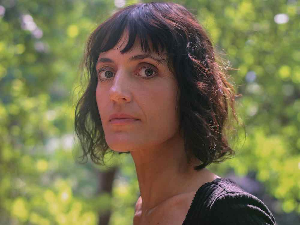 Astra Taylor looks directly at the camera with a knowing expression. She has wavy dark brown hair that cuts above her shoulders. She is wearing a black top and stands outdoors on a sunny day. In the background, bright green foliage is in soft focus.