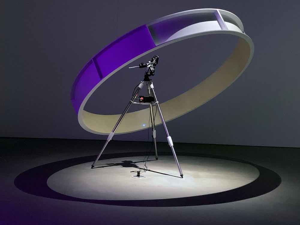 A purple circular contraption is mounted on a helix above a tripod mounted with small cameras. The items are standing on a grey floor in a dimly lit indoor studio space.
