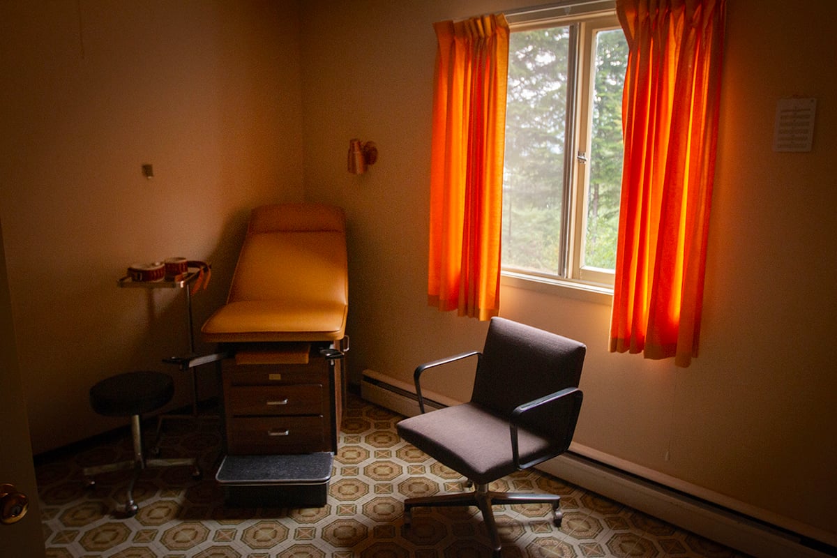 A dimly lit room with a swivel chair and examination table. Trees can be seen through a window with orange curtains.