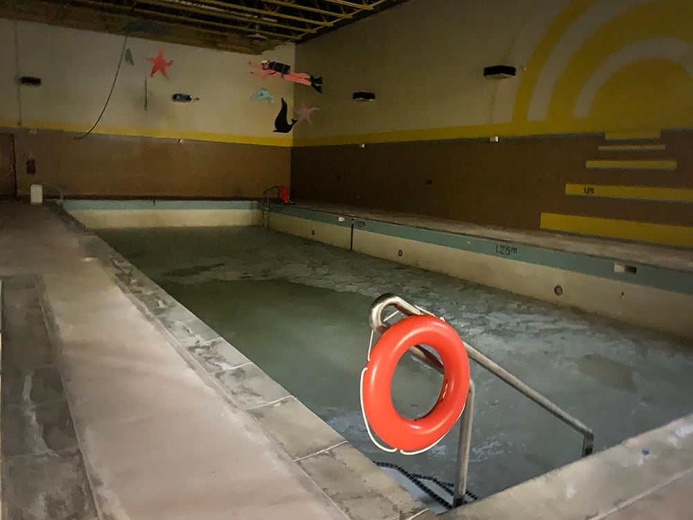An empty swimming pool sits in a darkened building. There is a red life-saving buoy hanging from the pool’s hand rail.