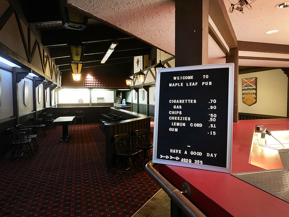 A darkened room with tables, chairs and a dartboard. A sign says, “Welcome to Maple Leaf Pub.” 