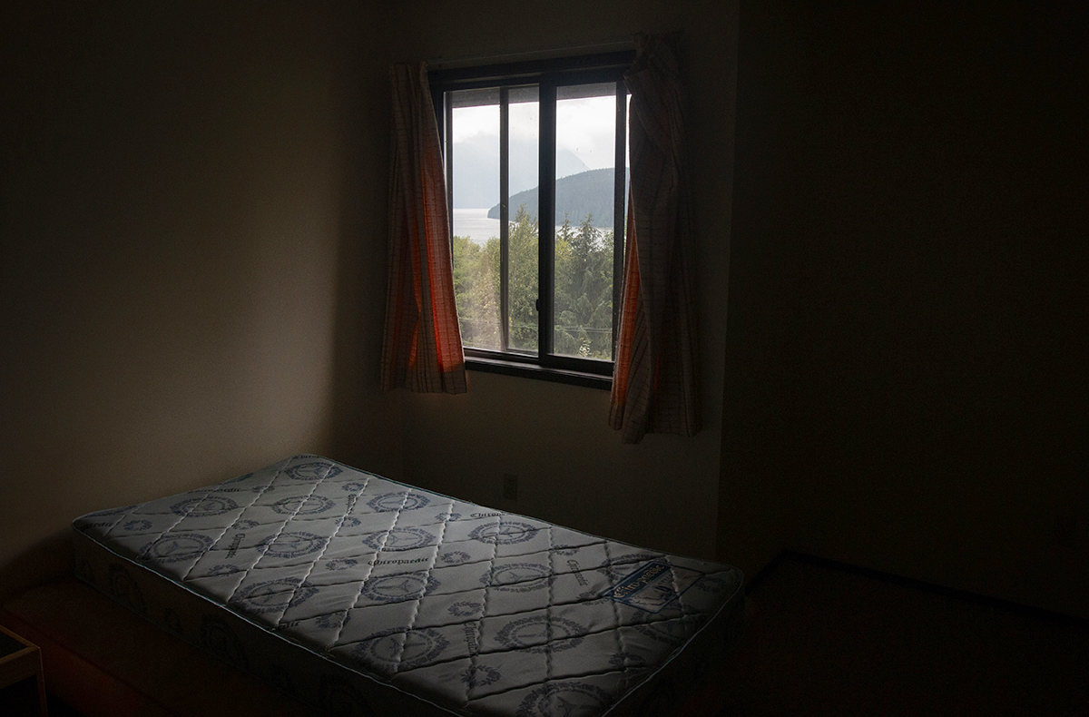 A darkened room with a bare mattress and a window with orange curtains. A coastal inlet can be seen out the window.