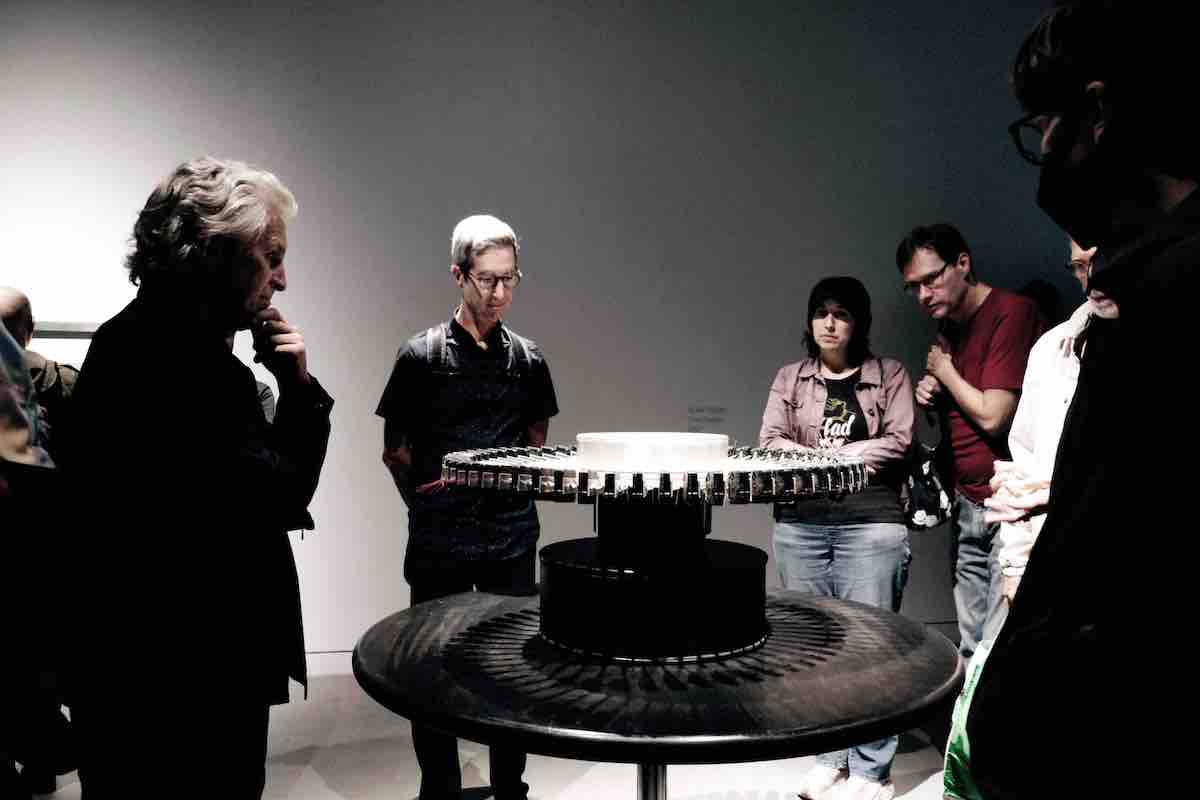 A group of people stand around a whirling circular contraption in a dimly lit studio space. The people, clad in casual streetwear, are all looking at the contraption.