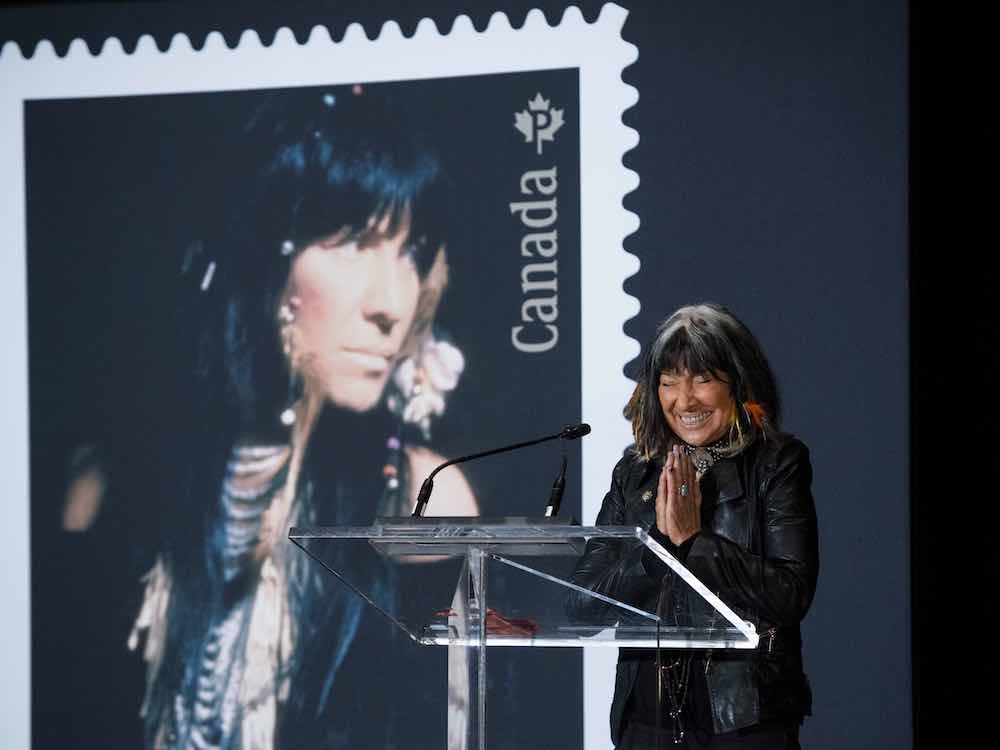 Buffy Sainte-Marie has dark shoulder-length hair, bangs and grey highlights. She holds her palms together in a gesture of gratitude while she smiles widely, closing her eyes. She is wearing dark clothing, including a black leather jacket, and stands at a glass podium in front of a large projection of a Canadian postage stamp bearing an archival portrait of her wearing Indigenous jewelry.