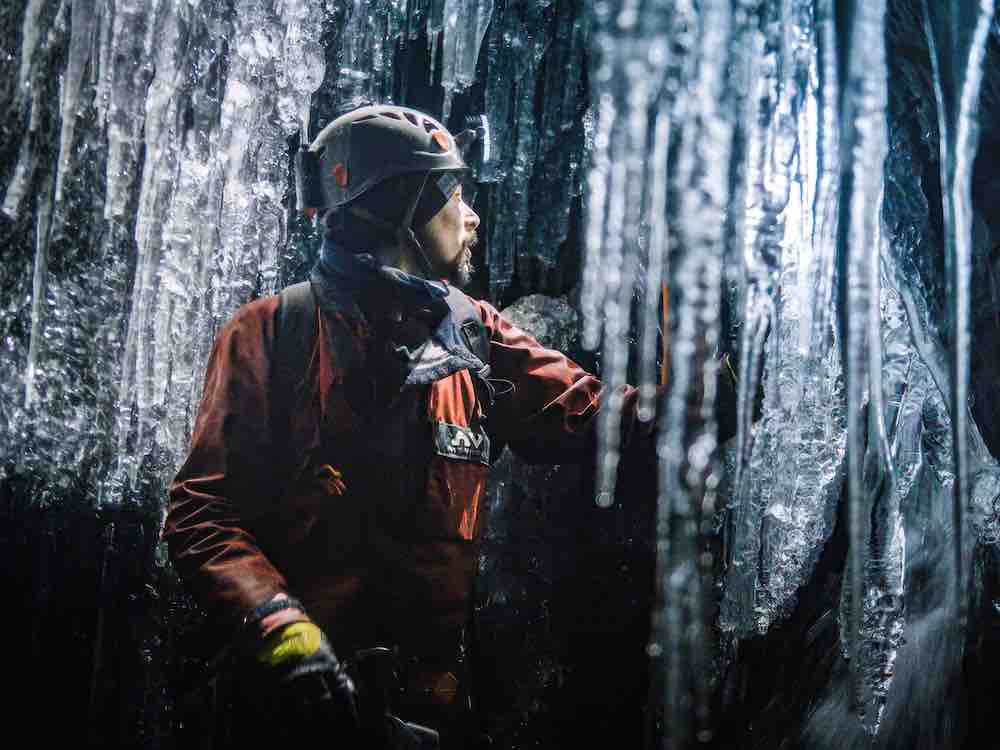 A man with a beard in a grey helmet and red jumpsuit looks towards the right of the frame, where his headlamp shines to illuminate a wall of icicles in a cave. The frame is filled with translucent ice against darkness.