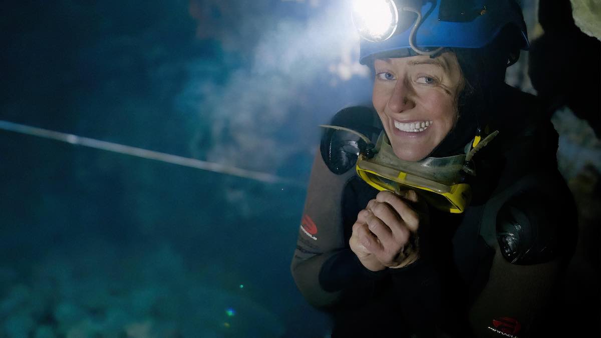 Katie Graham has blue eyes and is smiling widely at the camera. She stands to the right of the frame in a blue helmet with a headlamp, a pair of yellow diving goggles around her neck and a brown and black wetsuit to ready herself for diving. Beside and behind her is the murky deep blue depths of an underground pool, out of focus.