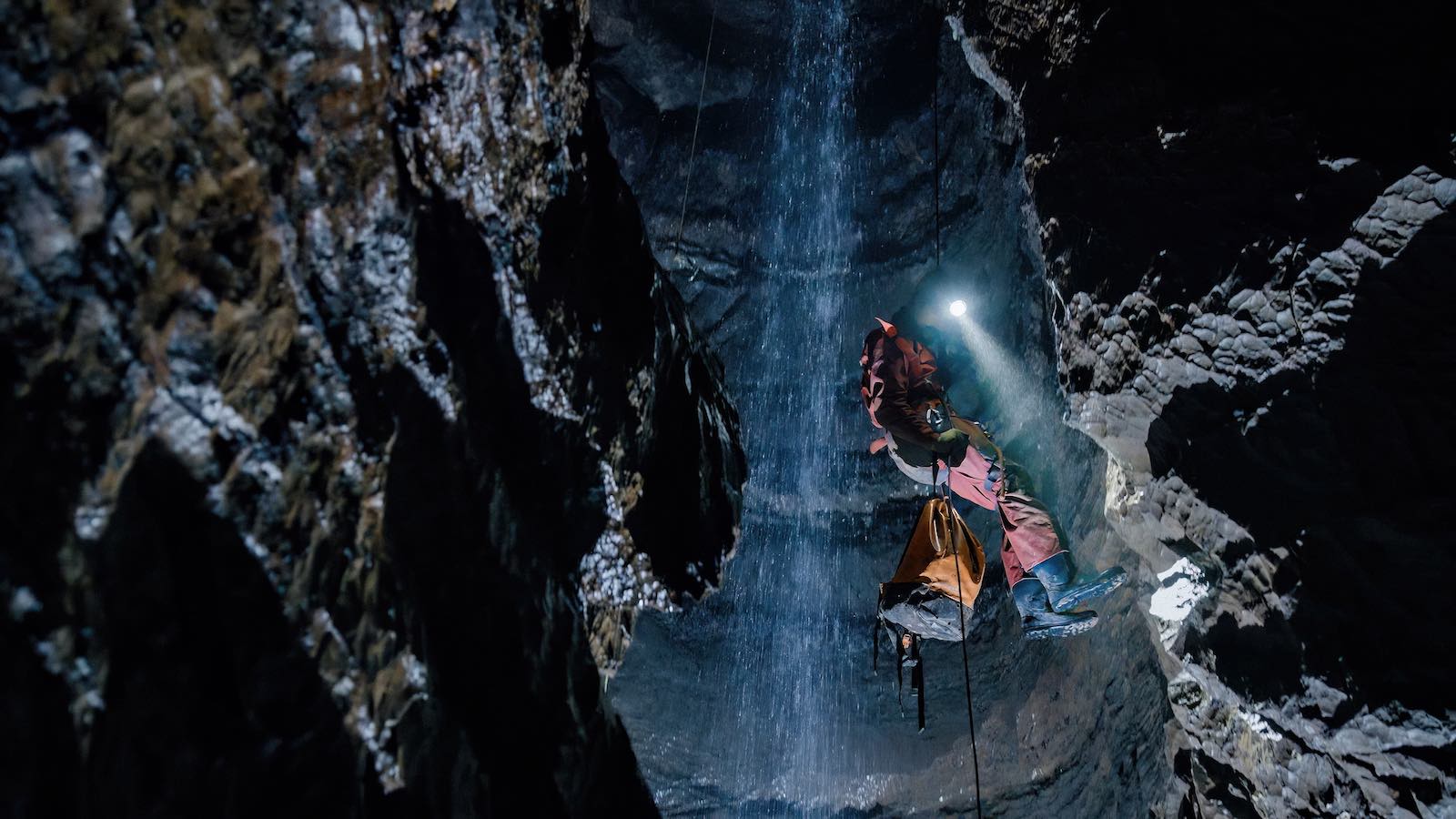 A person in a red jumpsuit and black calf-length boots rappels beside an underground waterfall. An orange rucksack is attached to their black ropes and their headlamp shines downward to the right of the frame. The person is framed by craggy vertical faces of dark rock on all sides.