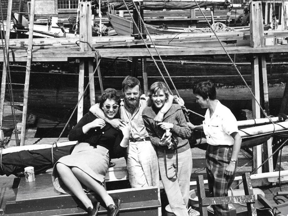 A black and white photo of four people on a wooden dock. Farley Mowat, second from left, has his arms around a French student, left, and Claire Mowat, right.