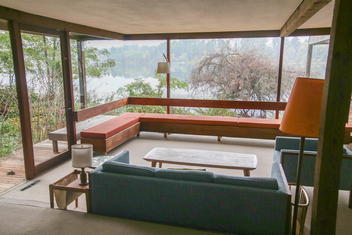 A mid-century playroom with a panoramic view of a lake on a cloudy day. There is an orange-cushioned bench that wraps around the glass windows in an L shape.
