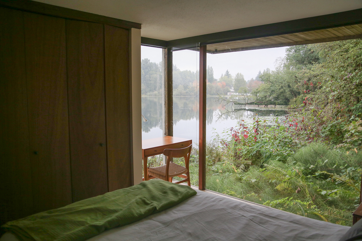 A bedroom with a window overlooking Deer Lake. There is a faux-teak desk with a Danish cord chair.