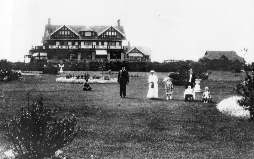 A black and white photo of an Arts and Crafts mansion with a family of six, and presumably a nanny, in period clothing.