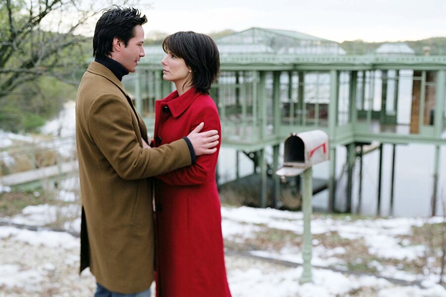 Keanu Reeves and Sandra Bullock hold each other in front of a lake house.