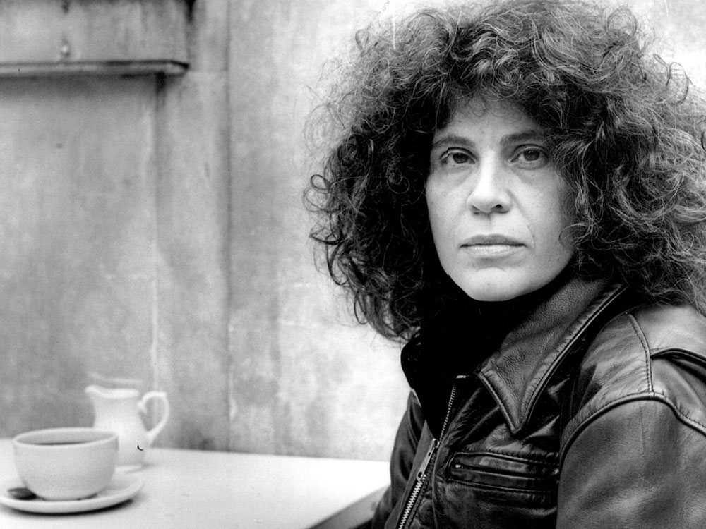 A black and white portrait of Anne Michaels depicts a woman with dark curly hair and a leather jacket sitting to the right of the frame at a table in a coffee shop. She is looking directly at the camera with a serious expression. On the table before her is a small coffee cup and small pitcher for cream.
