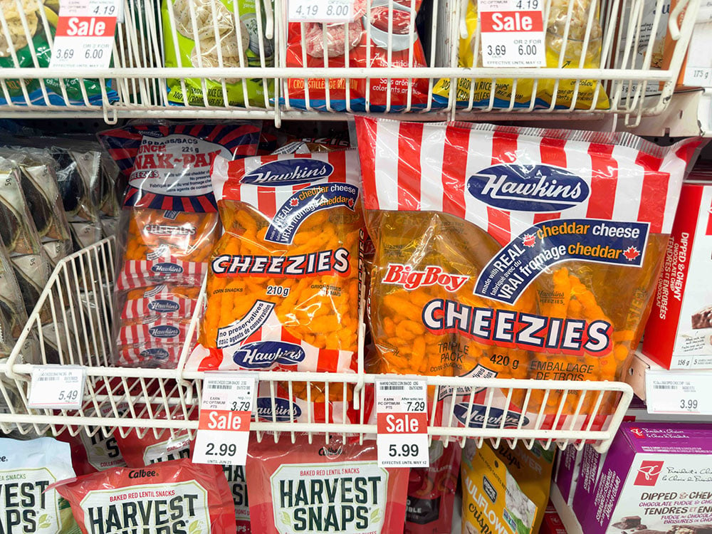 A Canadian drugstore shelf features bright orange bags of Hawkins Cheezies in distinctive red and white packaging. In the shelves above and below are other packaged snack foods.