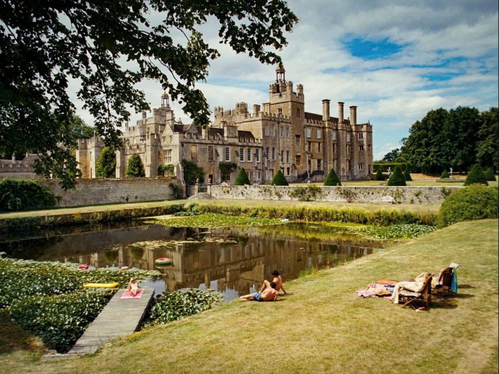 A portrait of a castle-like English manor on a sunny day. In the foreground, people lounge in bathing suits on the grass near the water.
