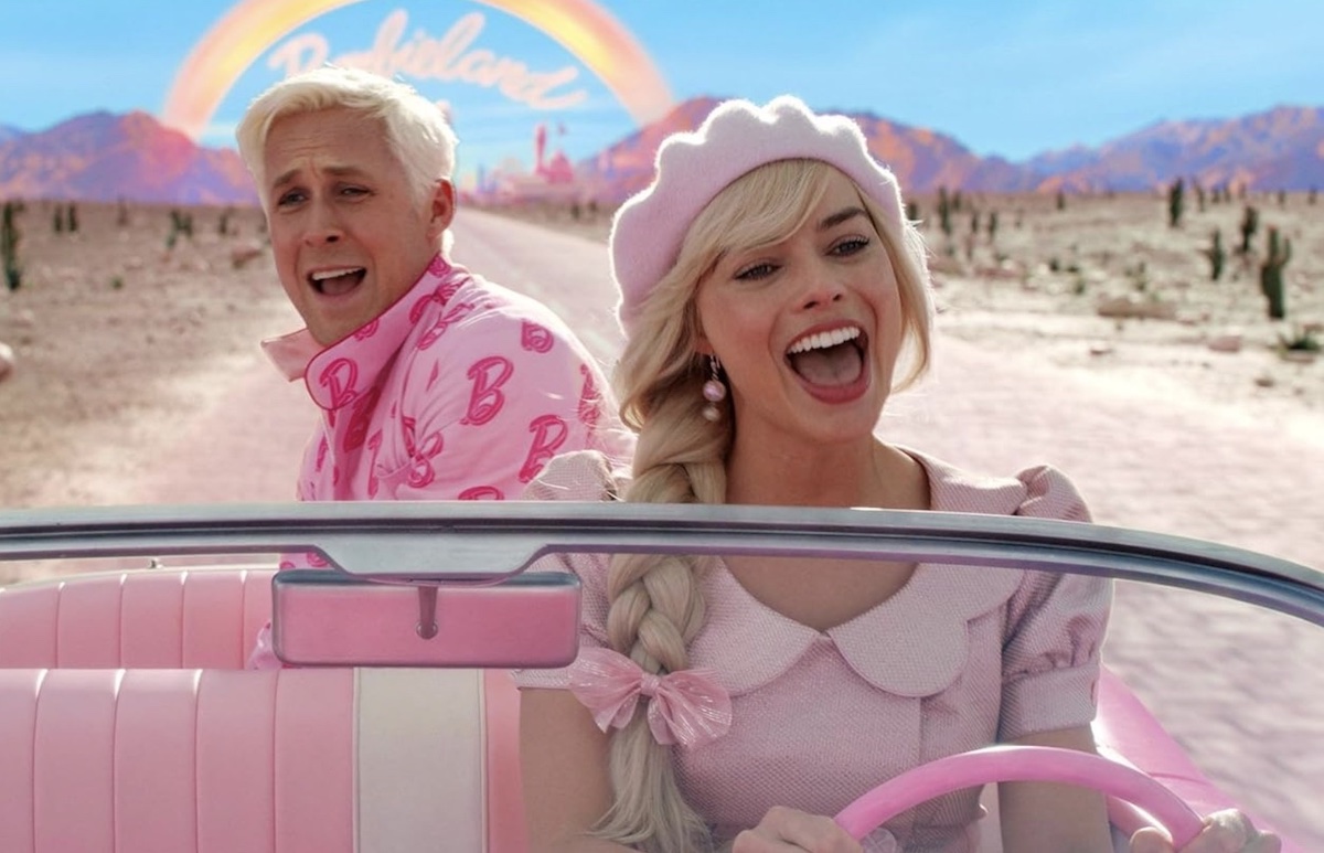 Two people ride a small pink convertible car through a desert with the roof rolled down. Behind them, a rainbow spans across a mountain range under which the word 'Barbieland' is faintly visible in stylized script. To the left, a man with short white-blond hair is in the back seat, smiling, singing and attired in bright pink. To the right, a woman is in the driver’s seat, also smiling and singing. Her long blond hair is in a thick braid; she is wearing a baby-pink beret and matching dress.