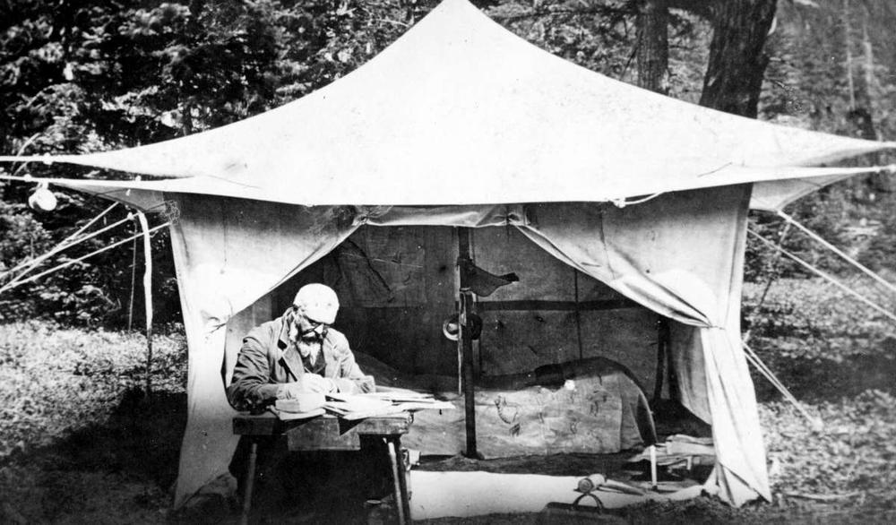 A black and white photo of an older man with a beard and glasses consulting a pile of paperwork in front of a canvas tent.