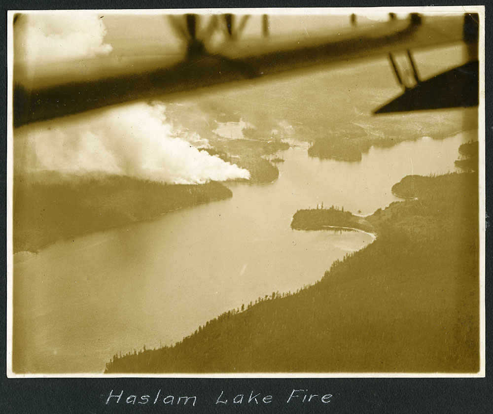 A sepia-toned photo reads 'Haslam Lake Fire.' The photo has been taken from a float plane — the wing is visible — as trees ringing Haslam Lake send up plumes of smoke.