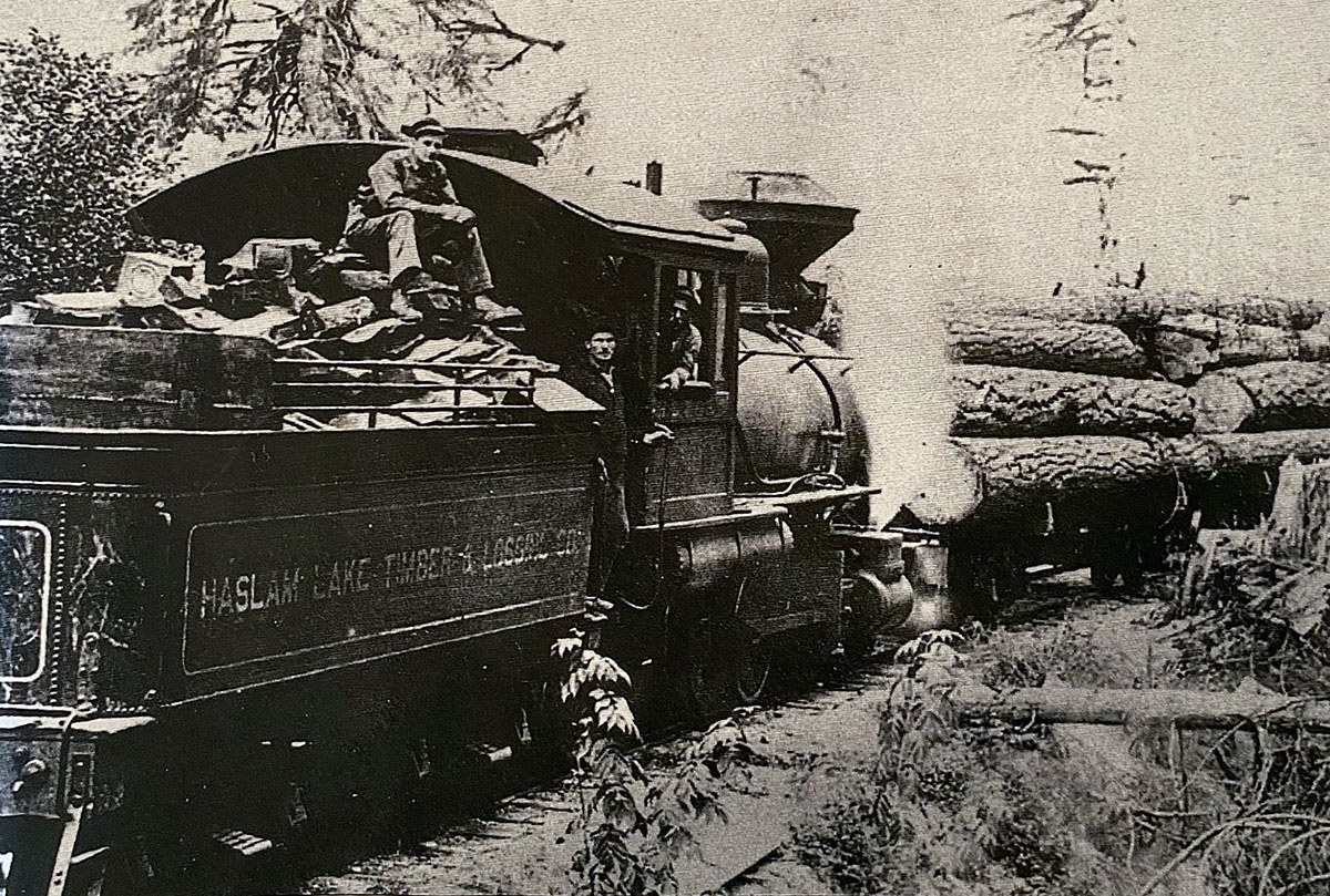 A black and white photo shows a steam locomotive hauling logs. A man sits on the logs that feed the engine; another staffs the locomotive, and a third poses between the other two men.