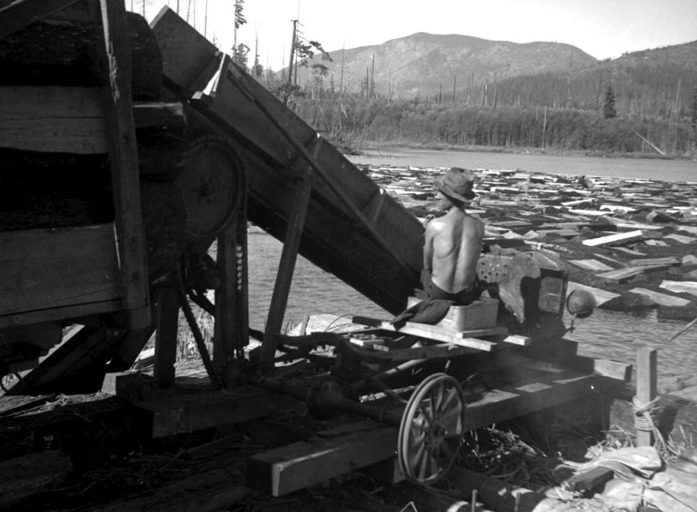 A black and white photo shows a shirtless man sitting and working a machine that is removing blocks of wood from Haslam Lake.