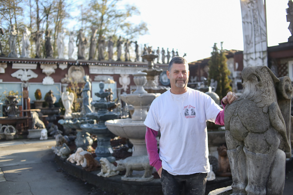 A man stands with his hand on a griffin in a yard full of statues.