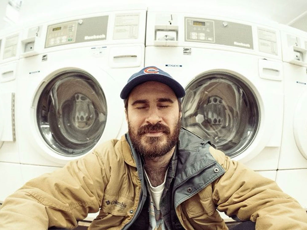 A man with long brown hair, a beard and a blue Chicago Cubs ball cap wears a beige jacket over a white T-shirt. He sits with his eyes closed on the floor of a laundromat. Behind him is a row of white washing machines under fluorescent lights.