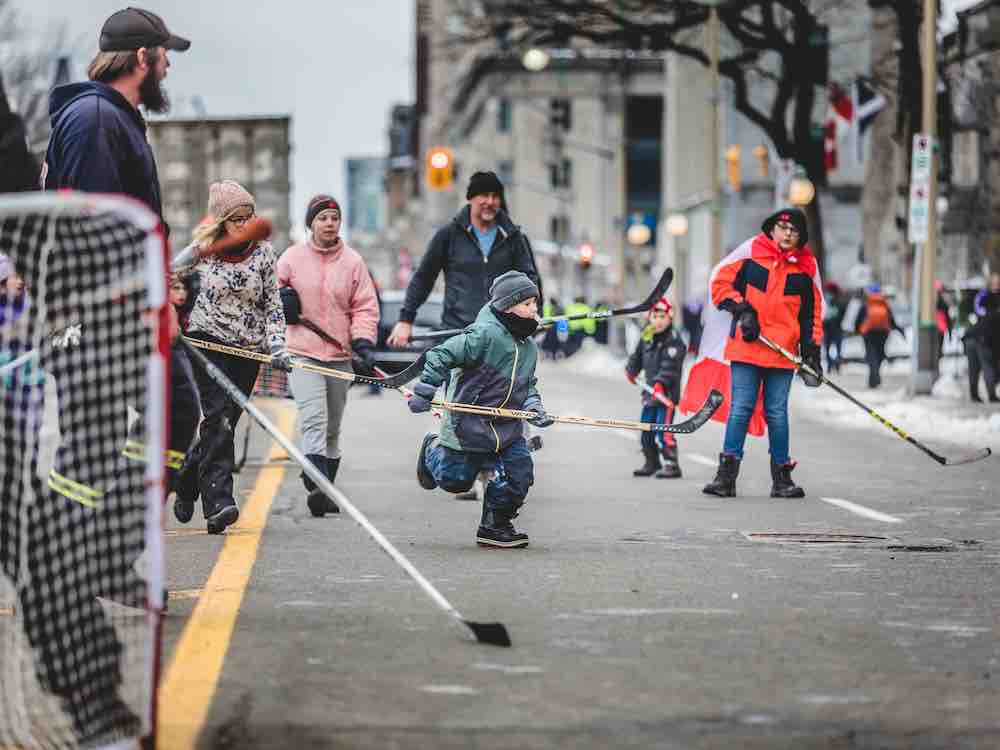 A group of kids and adults play road hockey on the street. All appear to be light-skinned and are wearing winter clothing. Many are adorned with a piece of clothing bearing a maple leaf. One child wears a Canada flag as a cape.