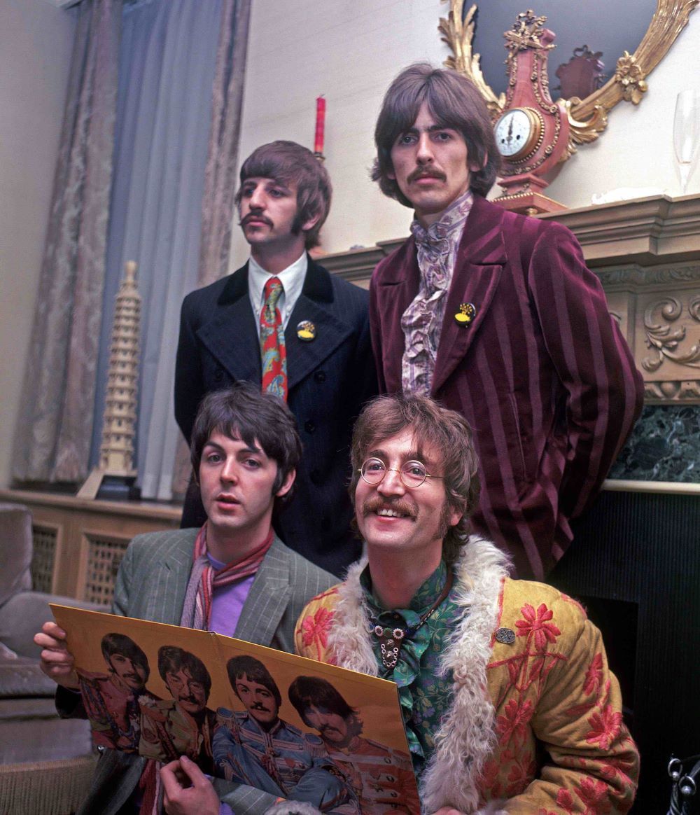 A colour archival photograph of four men in jewel-toned suits standing in front of an ornate mantel. The two men seated in front of those standing behind them are holding open a vinyl record cover featuring the four of them in colourful outfits against a yellow background. It is the interior spread of their album 'Sgt. Pepper’s Lonely Hearts Club Band.'