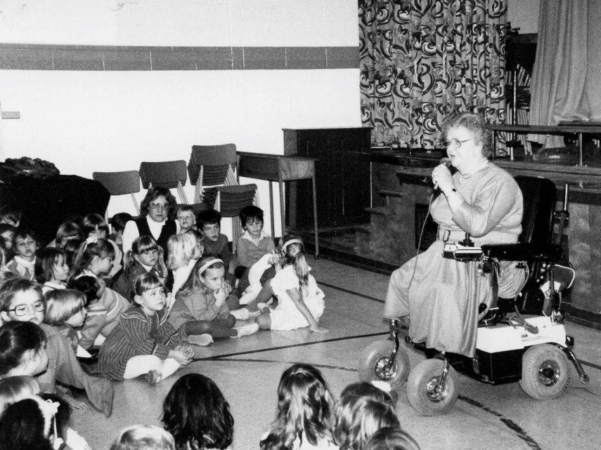 A black and white photo of a light-skinned woman with short, curly hair and glasses, using a motorized wheelchair and holding a microphone. She is in a school gymnasium. Elementary school kids sit on the floor, with some teachers among them, listening.