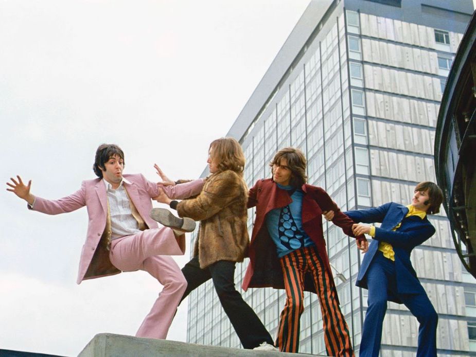 Four men pose atop the concrete roof of a building. On the left, a man in a pink suit with a white shirt is pretending to lose his balance while those to the right gesture to pull him towards them to safety. From left, the men next to the man in the pink suit are wearing a brown fur coat, a long burgundy coat and striped pants, and a blue suit over a yellow shirt.