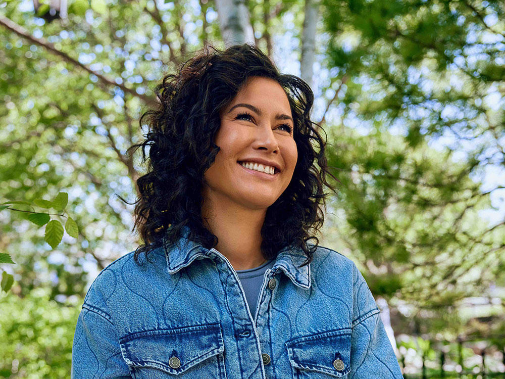 Sarika Cullis-Suzuki has olive skin and dark curly shoulder-length hair. She is looking up towards the horizon, smiling. She wears a light blue denim jacket over a blue top. In the background in soft focus is a stand of bright green deciduous trees against a blue sky.